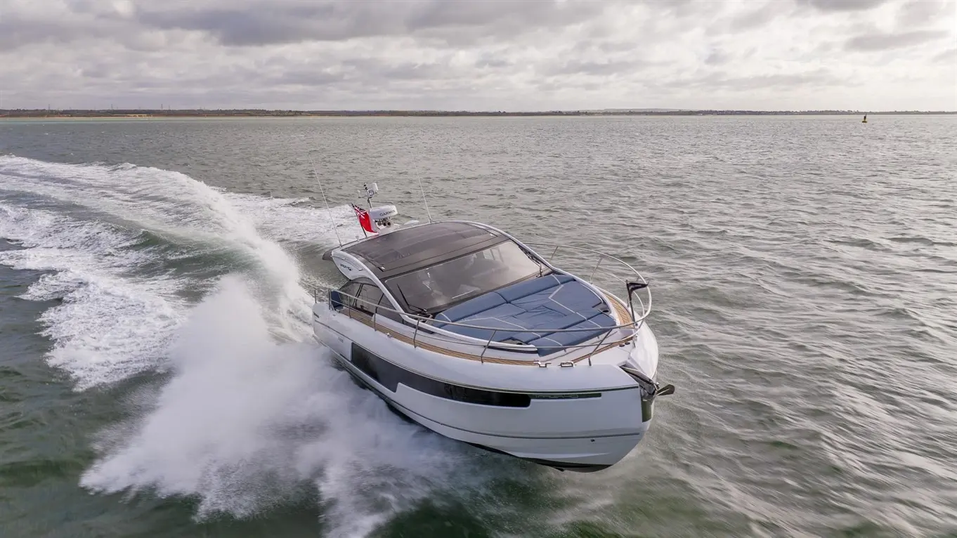 Luxury motorboat gliding over water, creating a spray, with a red flag flying and cloudy skies in the background.